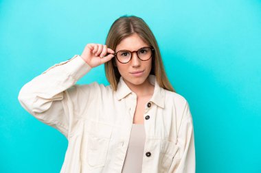 Mavi arka planda izole edilmiş genç beyaz bir kadın gözlüklü ve sinirli bir ifadeyle