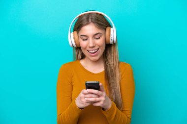 Genç beyaz kadın mavi arka planda izole edilmiş müzik dinliyor ve cep telefonu arıyor.