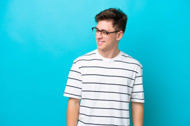 Genç ve yakışıklı Brezilyalı bir adam mavi arka planda izole edilmiş, yana bakıyor ve gülümsüyor.