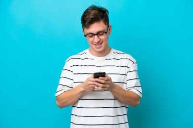 Young handsome Brazilian man isolated on blue background sending a message with the mobile