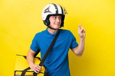 Young Brazilian man with thermal backpack isolated on yellow background with fingers crossing and wishing the best