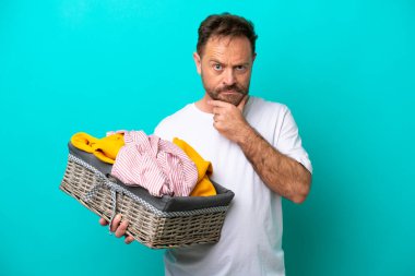Middle age woman holding a clothes basket isolated on blue background thinking