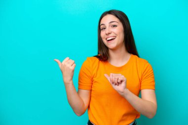 Mavi arka planda izole edilmiş genç beyaz kadın bir ürünü sunmak için kenarı işaret ediyor.