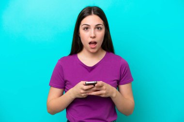 Young girl isolated on blue background looking at the camera while using the mobile with surprised expression