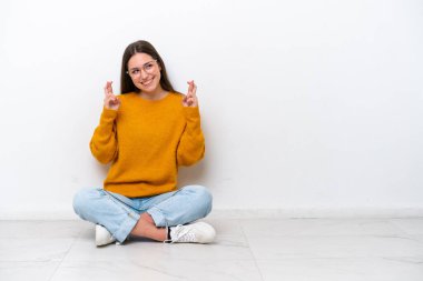 Young girl sitting on the floor isolated on white background with fingers crossing