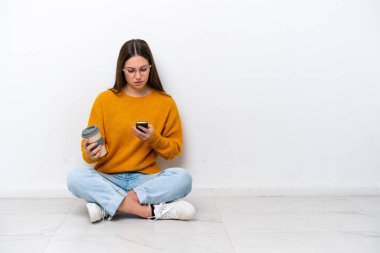 Young girl sitting on the floor isolated on white background holding coffee to take away and a mobile