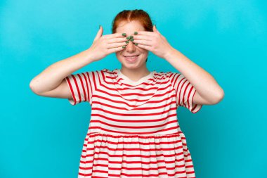 Young reddish woman isolated on blue background covering eyes by hands and smiling