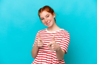 Young reddish woman isolated on blue background pointing to the front and smiling