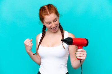 Young caucasian reddish holding a hairdryer isolated on blue background celebrating a victory