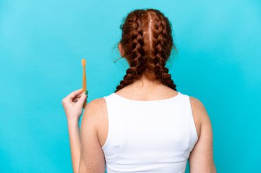 Young caucasian reddish woman brushing teeth isolated on blue background in back position