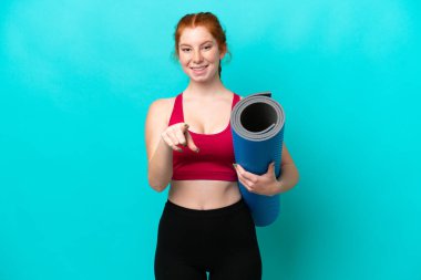 Young sport reddish woman going to yoga classes while holding a mat isolated on blue background surprised and pointing front