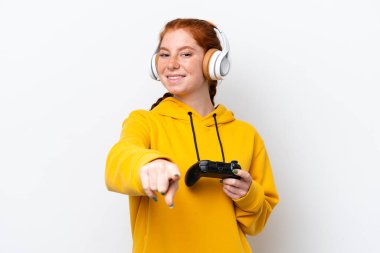 Young reddish woman playing with a video game controller isolated on white background pointing front with happy expression