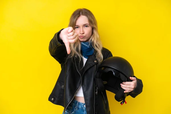 Blonde English young girl with a motorcycle helmet isolated on yellow background showing thumb down with negative expression