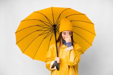 Teenager girl with rainproof coat and umbrella over isolated white background with tired and sick expression