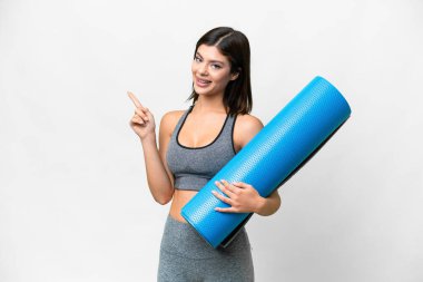 Young sport woman going to yoga classes while holding a mat over isolated white background pointing to the side to present a product clipart