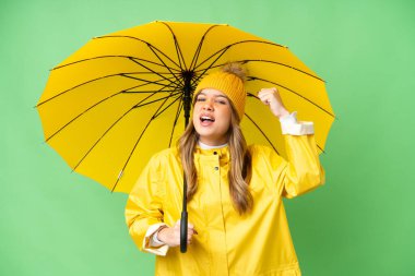 Yağmura dayanıklı paltosu ve şemsiyesi olan genç bir kız izole edilmiş krom anahtar arka planda zaferi kutluyor.