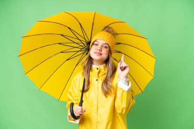 Yağmura dayanıklı paltosu ve şemsiyesi olan genç bir kız izole edilmiş krom anahtar arka planda en iyinin işareti olarak parmağını kaldırıyor ve gösteriyor.