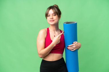 Genç sporcu kız yoga derslerine giderken izole edilmiş krom anahtar arkaplanının üzerinde bir minder tutuyor ve bir ürünü sunmak için yan tarafı işaret ediyor.