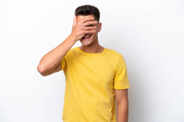 Beyaz arka planda izole edilmiş beyaz tenli genç bir adam. Gözleri elleriyle kaplıyor ve gülümsüyor.