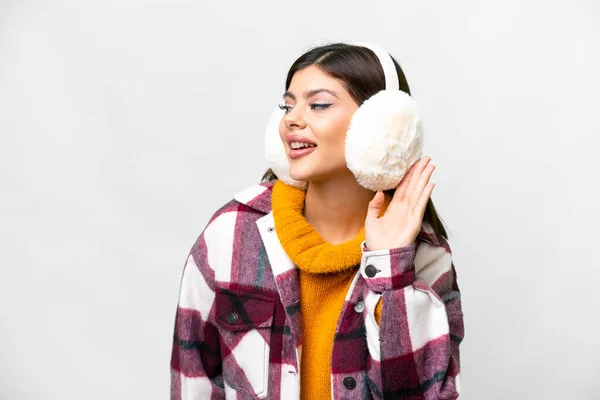 Young Russian woman wearing winter muffs over isolated white background listening to something by putting hand on the ear