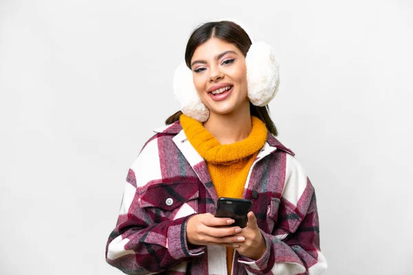 Young Russian woman wearing winter muffs over isolated white background sending a message with the mobile