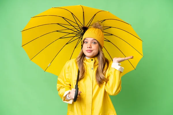 若いです女の子とともにレインプルーフコートと傘上の隔離されたクロマキーバック持っています疑問ながら手を上げます — ストック写真