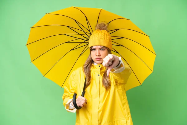 若いです女の子とともにレインプルーフコートと傘上の隔離されたクロマキー背景ショー親指ダウンとともに負の式 — ストック写真