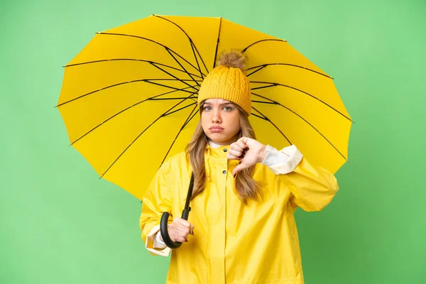 穿着防雨外套 披着雨伞的小女孩 背景是孤立的色 带着负面表情 大拇指垂下 — 图库照片