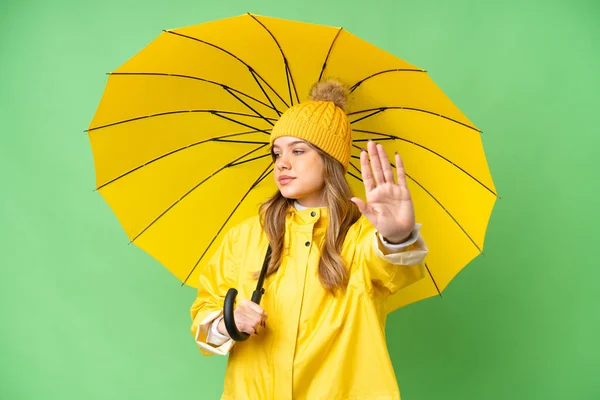 穿着雨衣 披着雨伞的小女孩在孤零零的色彩关键的背景上做着停止的手势和失望 — 图库照片