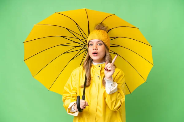 年轻姑娘穿着防雨外套 带着雨伞 遮挡着孤立的彩色关键背景 想在举起一只手指头的同时实现这个解决方法 — 图库照片