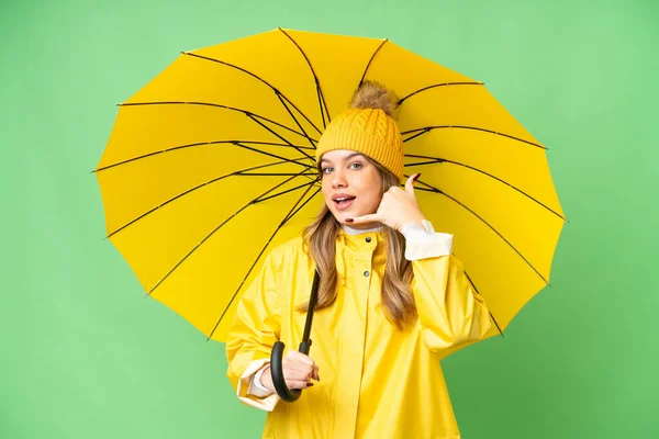 携帯電話のジェスチャーを作る隔離されたクロマキーの背景の上にレインプルーフコートと傘を持つ若い女の子 サインを呼んでくれ — ストック写真