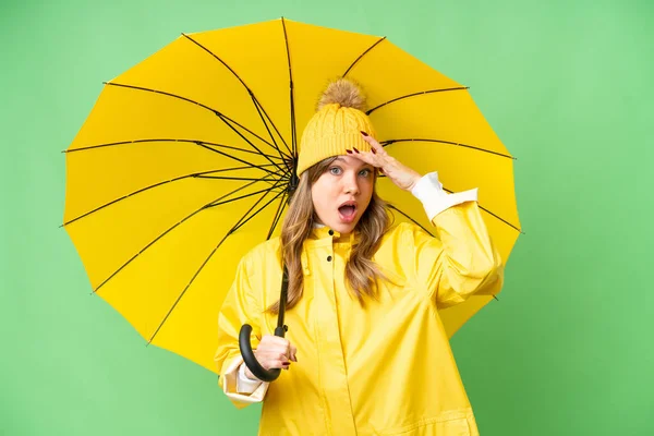 年轻姑娘穿着防雨外套 披着雨伞 在孤立的彩色关键背景上做着令人惊讶的手势 同时向旁边望去 — 图库照片