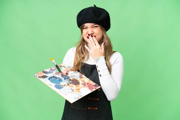 年轻的艺术家女孩手牵着调色板 在孤立的色彩键背景上欢快地微笑着 用手捂着嘴 — 图库照片
