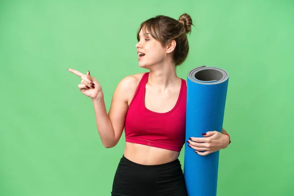 年轻的女运动员一边去上瑜伽课 一边拿着一个垫子盖着孤立的彩色关键背景 手指指向旁边 并展示一种产品 — 图库照片