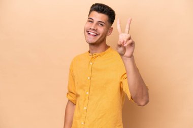 Beyazlı genç adam bej arka planda izole edilmiş gülümsüyor ve zafer işareti gösteriyor.