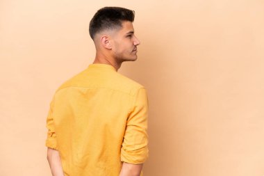 Young caucasian man isolated on beige background in back position and looking back