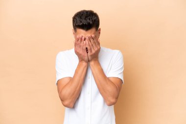 Beyazlı genç bir adam bej arka planda izole edilmiş, yorgun ve hasta bir ifadeyle.