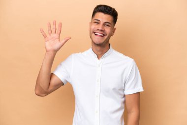 Beyazlı genç bir adam bej arka planda izole edilmiş mutlu bir ifadeyle selamlıyor.