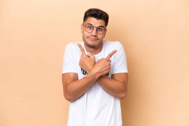 Young volunteer caucasian man isolated on beige background pointing to the laterals having doubts