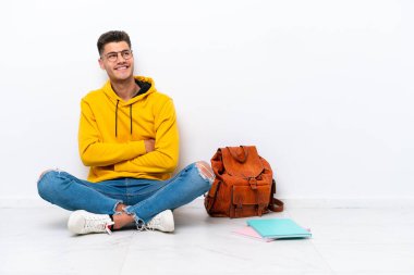 Young student caucasian man sitting one the floor isolated on white background looking up while smiling