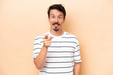 Beyazlı genç adam bej arka planda izole edilmiş şaşırmış ve ön tarafı işaret ediyor.