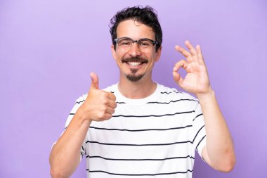Mor arka planda gözlüklü ve iyi bir işaret veren beyaz bir genç.
