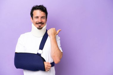 Mor arka planda izole edilmiş askı ve boyunluk takan genç beyaz bir adam bir ürün sunmak için yan tarafı işaret ediyor.