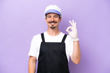 Fishmonger man wearing an apron isolated on purple background showing ok sign with fingers