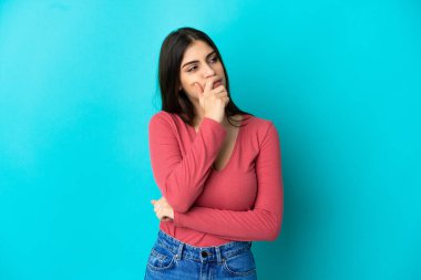 Kafkasyalı genç bir kadın mavi arka planda izole edilmiş. Şüpheleri var ve kafası karışmış bir yüz ifadesi var.