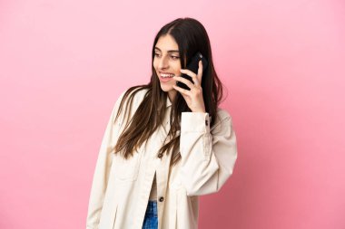 Pembe arka planda izole edilmiş genç beyaz bir kadın cep telefonuyla sohbet ediyor.