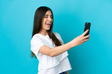 Mavi arka planda izole edilmiş beyaz bir kadın cep telefonuyla selfie çekiyor.