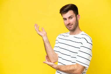 Sarı arka planda izole edilmiş genç beyaz adam davet etmek için ellerini yana uzatıyor.