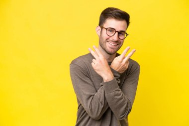 Sarı arka planda izole edilmiş genç beyaz adam gülümsüyor ve zafer işareti gösteriyor.