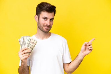 Young caucasian man taking a lot of money isolated on yellow background pointing finger to the side